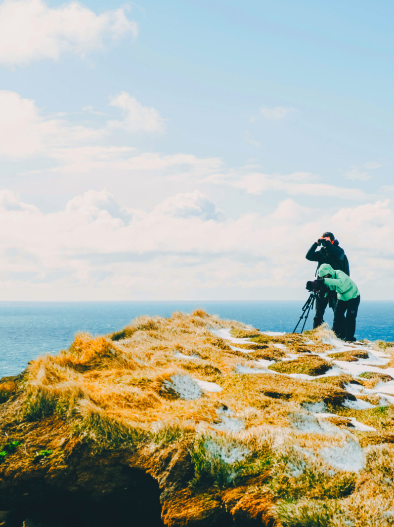 Two photographers taking pictures on a cliff against the ocean. Latrabjarg, Iceland