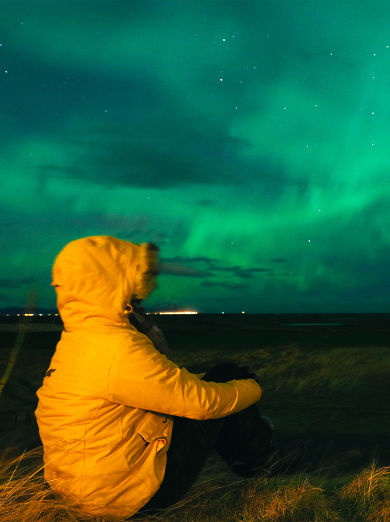 woman wearing a bright yellow winter jacket is sitting in a field as she is watching a Northern Lights display during a strong aurora storm in Southern Iceland in November 2015