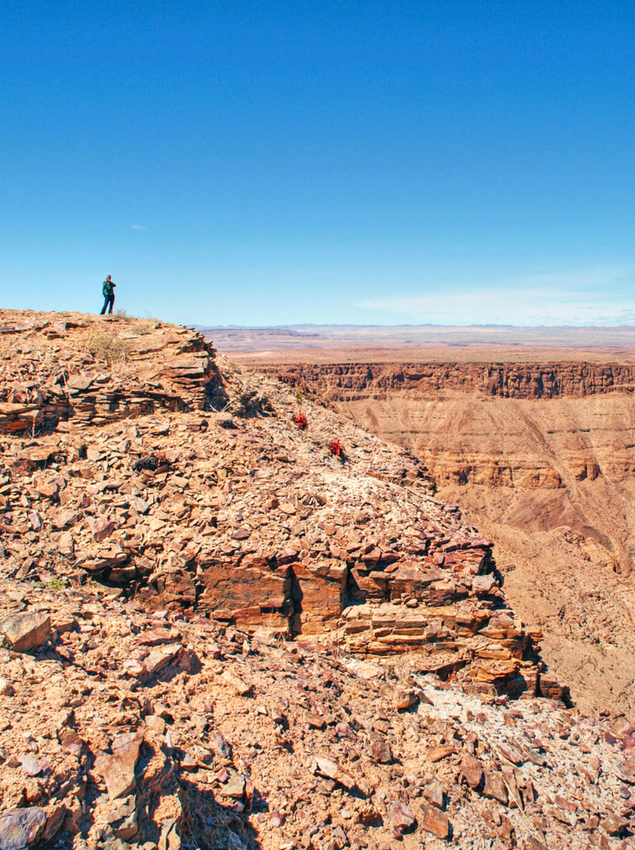 A woman stands upon a rocky precipice admiring a dramatic canyon view over Fish River Canyon, Namibia, Africa.