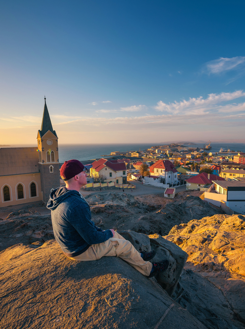 Tourist on top of a hill enjoys the panoramic view of Luderitz in Namibia at sunset, with lutheran church and atlantic ocean in the background.