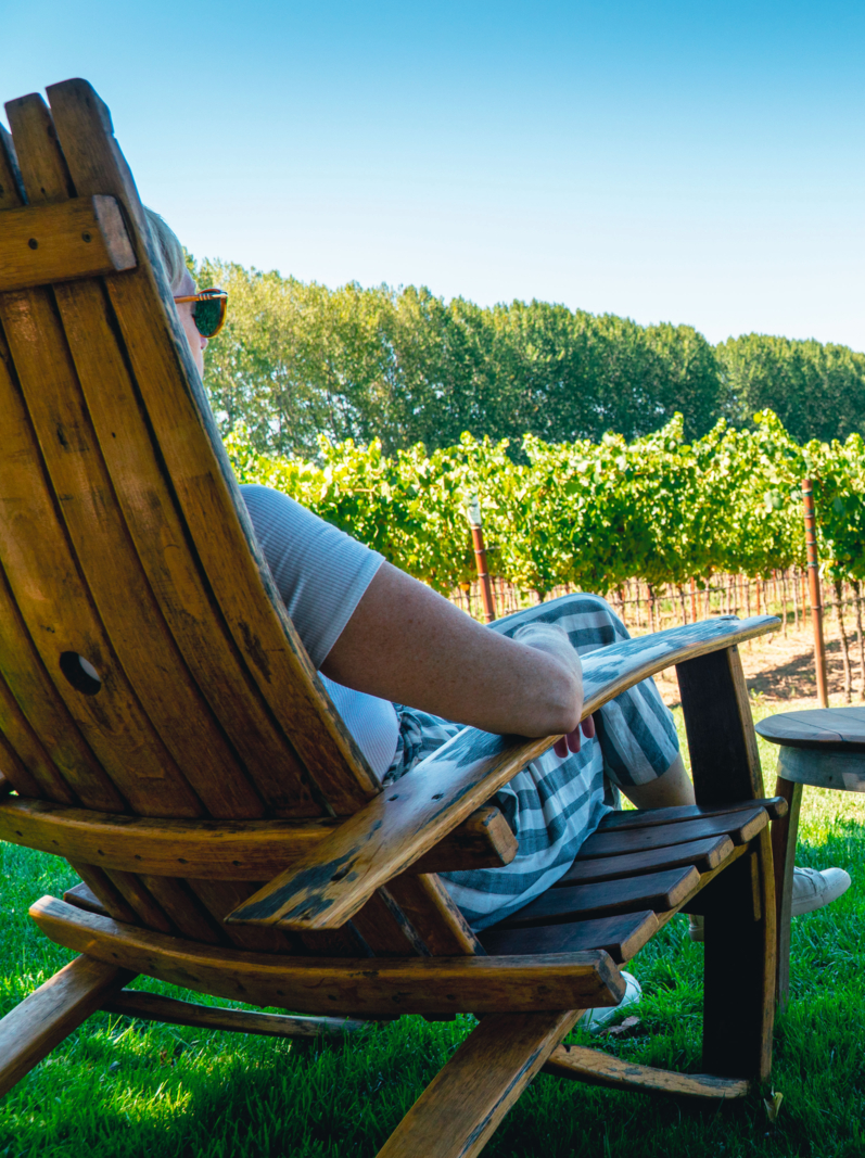 Middle aged woman sitting in a wooden chair while the seat next to her remains empty. She is looking at the beautiful view of the vineyard.