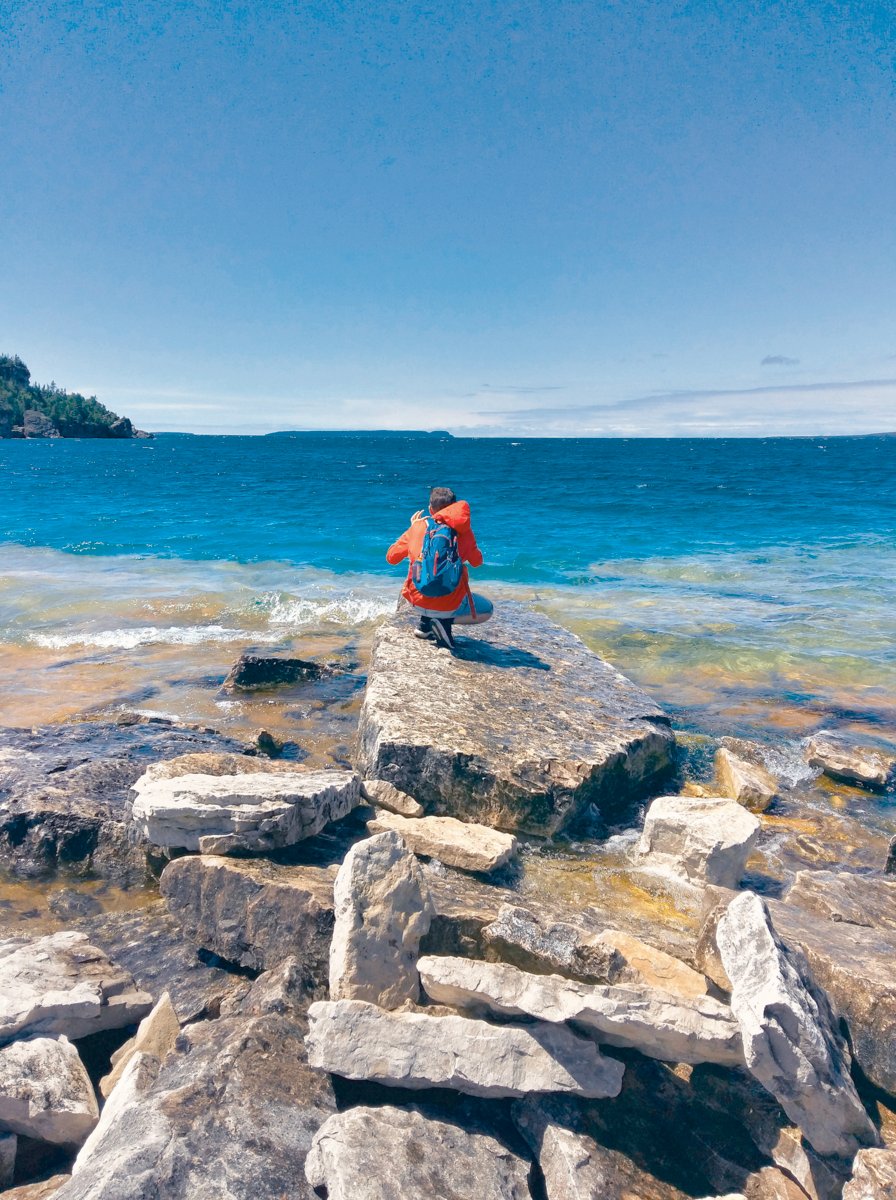 Young man in bright red jacket sitting on rocks at shore, taking photo of stunning seascape. Sunny day, clear sky, see thru turquoise water. Bruce Peninsula National Park, Tobermory, Ontario, Canada