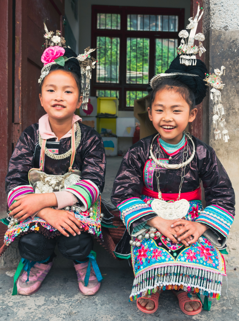 Two chinese dong people school girls sitting together in front of the entrance to their school classroom in traditional clothing in the small Dong Village Huang Gang
