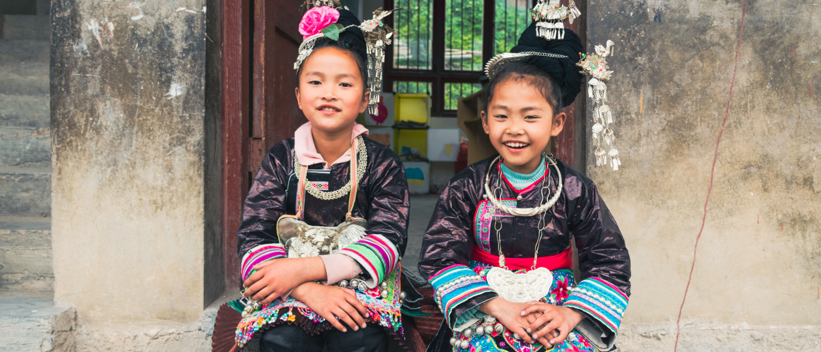Two chinese dong people school girls sitting together in front of the entrance to their school classroom in traditional clothing in the small Dong Village Huang Gang