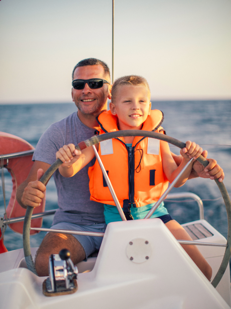 One little boy on sailboat sitting with his father, enjoying at summer and having a fun while steering with yacht at sunset