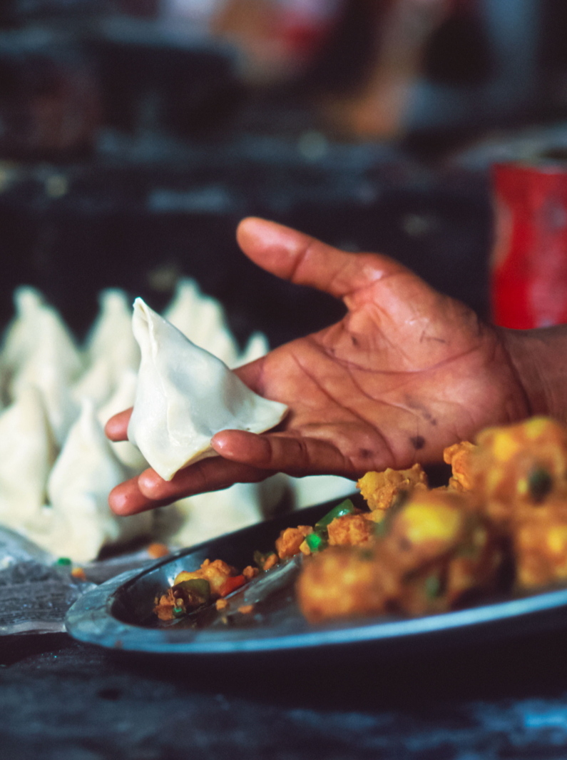 Horizontal image of a male hand holding a samosa ready for cooking and in the blurred background other numerous pieces resting on sheets of newspaper, traditional street food