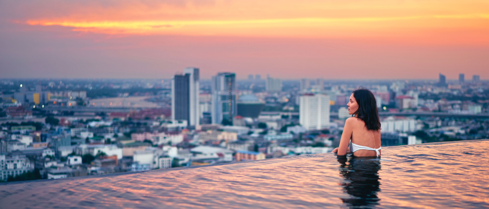 Young woman relax in swimming pool on roof top during amazing sunset and enjoy city view. Summer vacation and holidays concept