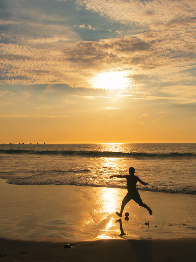 Young man playing soccer on the beach at sunset with sun shine reflecting in the sea