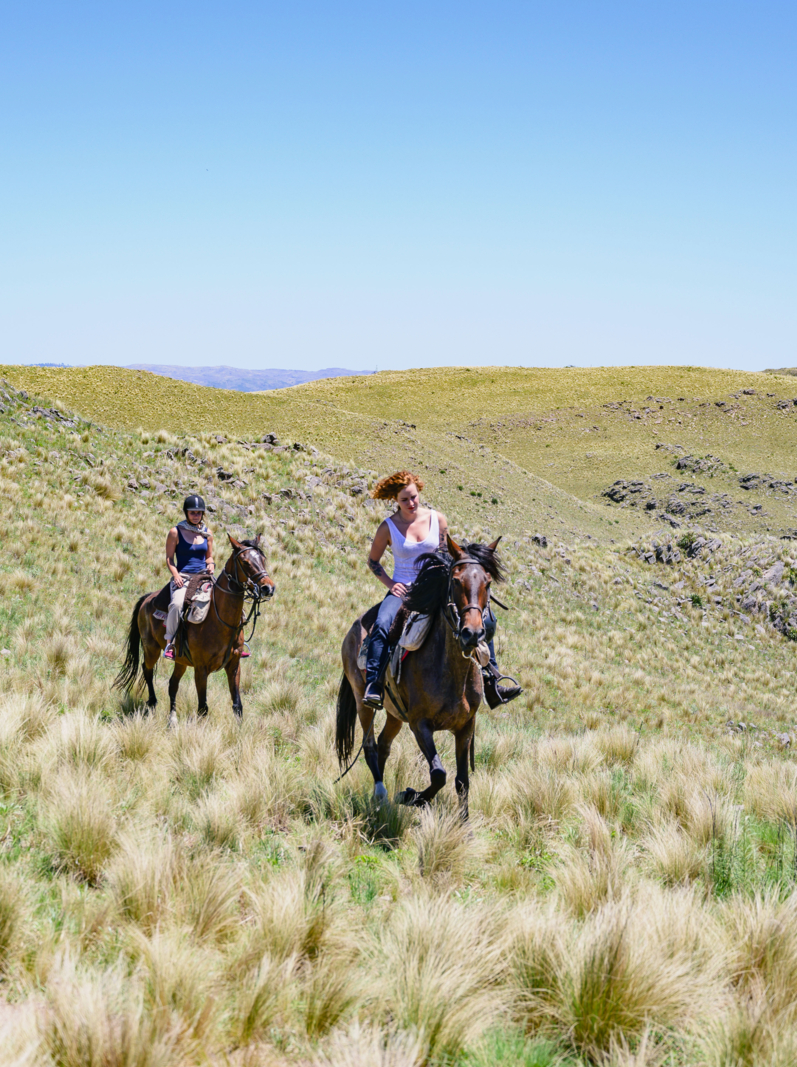 Two women in their 20s enjoying horseback riding through rocky outcrops and open hills in Argentine pampas.