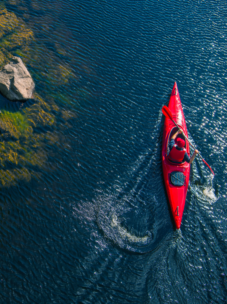 River Kayaker Aerial View. Caucasian Sportsman in the Red Kayak Paddling on the Scenic River Along the Shore